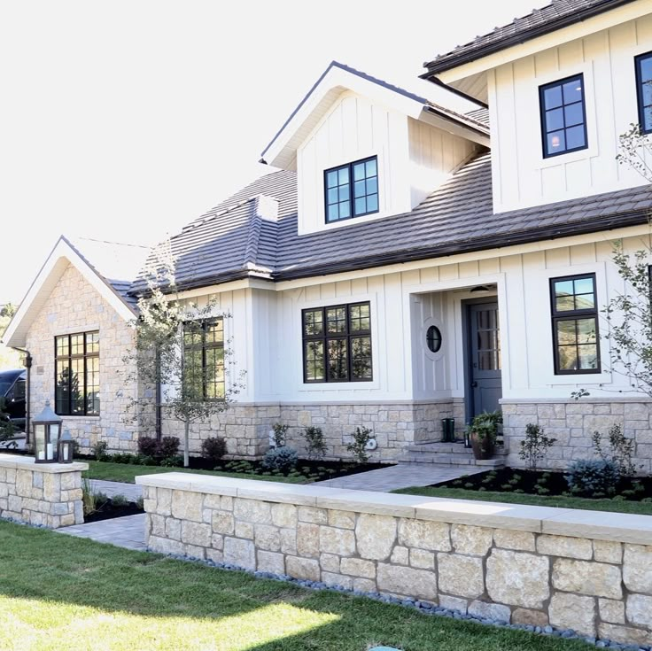 a white house with black windows and landscaping in the front yard, next to a stone wall