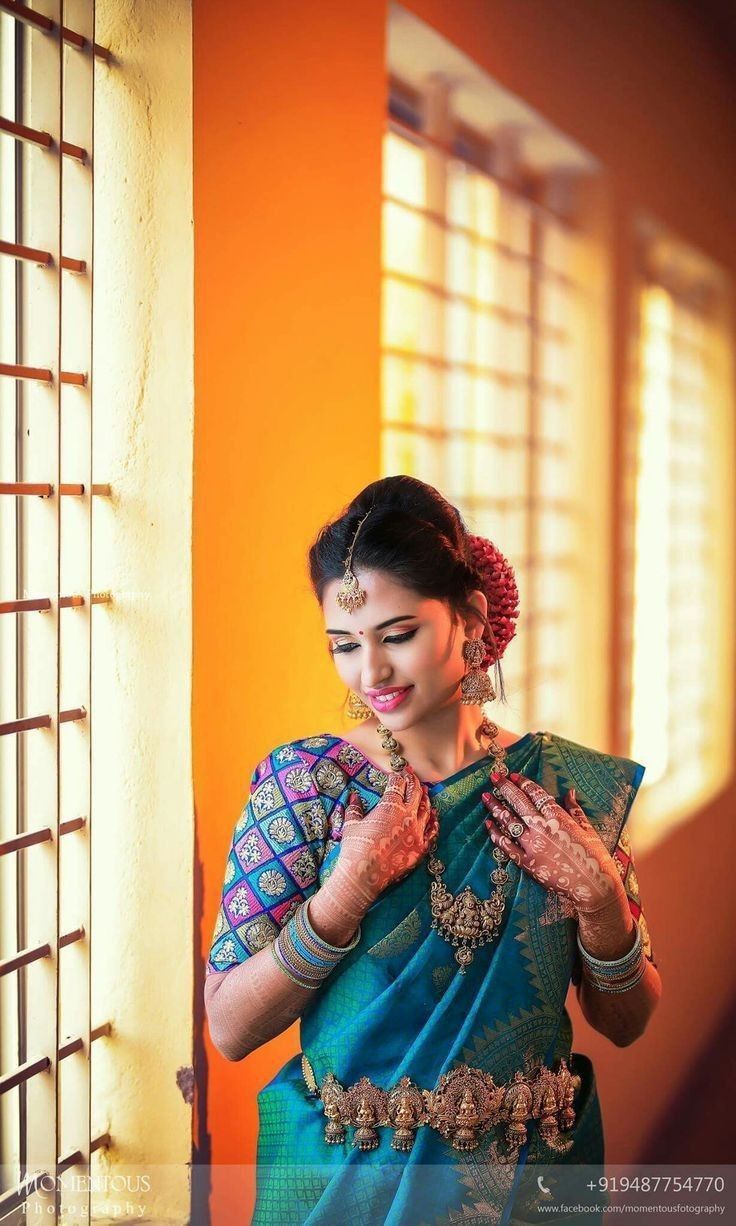 a woman in a blue and green sari standing by a window with her hands clasped to her chest