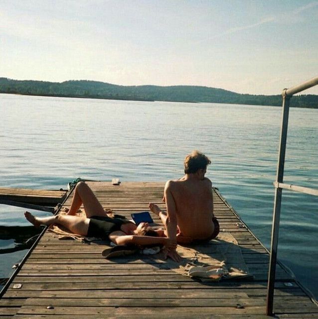 two people are sitting on a dock by the water and one is reading a book