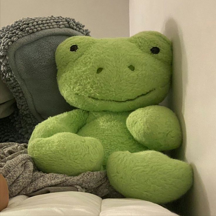 a green frog stuffed animal sitting on top of a bed next to a pillow and blanket