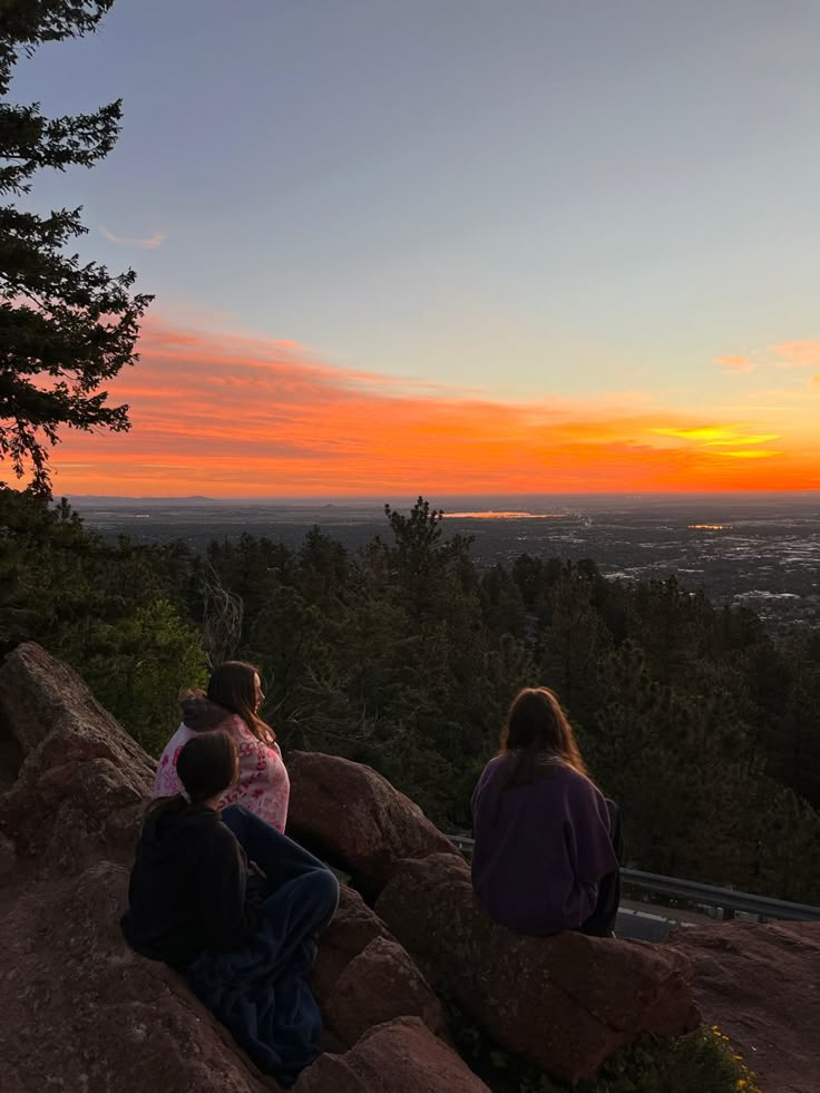 three people sitting on rocks looking at the sunset over a valley and city in the distance