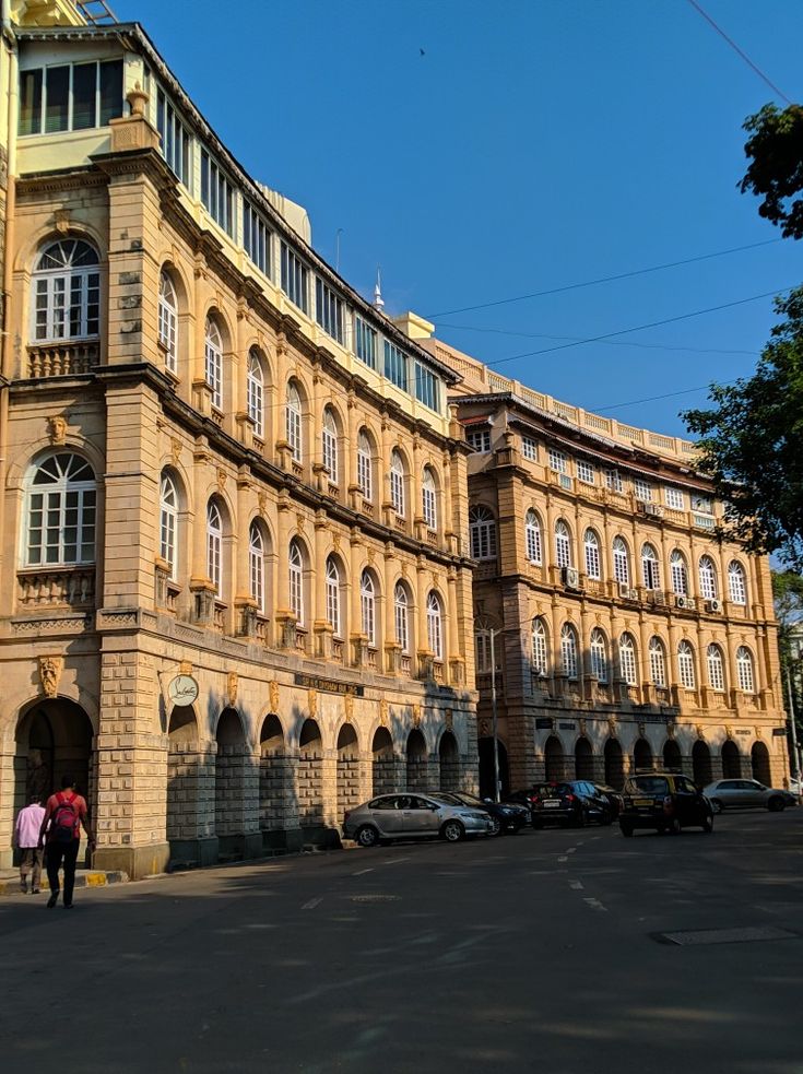 two people walking in front of an old building with cars parked outside and on the street