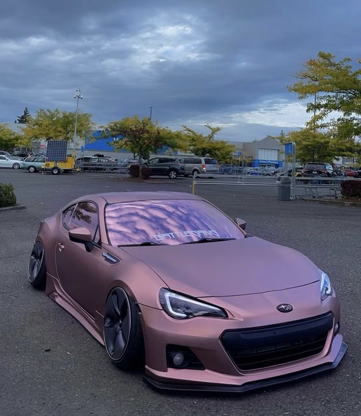 a pink sports car parked in a parking lot next to some bushes and trees on a cloudy day