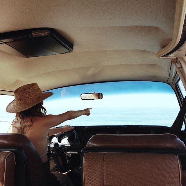 a woman wearing a hat sitting in the passenger seat of a car looking out at the ocean