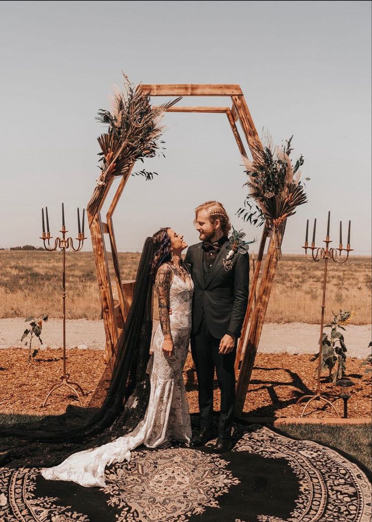 a man and woman standing next to each other in front of a wooden arch with greenery