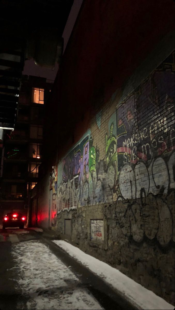 a wall covered in graffiti next to a red car on a snowy street at night