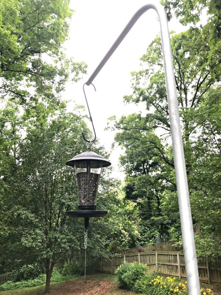 a bird feeder hanging from the side of a metal pole next to trees and flowers