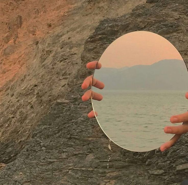 a person holding up a round mirror in front of a cliff by the water's edge