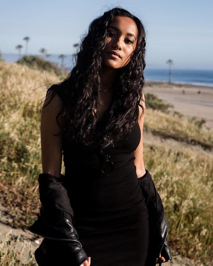 a woman in black dress and gloves standing by the beach with her hand on her hip