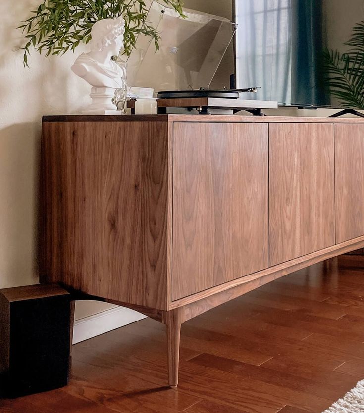 a wooden sideboard with a plant on top and a mirror in the corner next to it