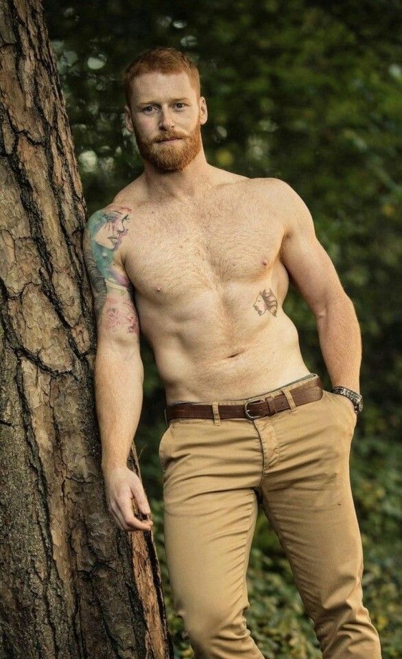 a shirtless man standing next to a tree in the woods with his hands on his hips