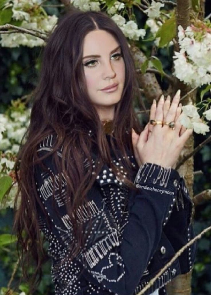 a woman with long hair standing in front of some flowers and holding her hands together