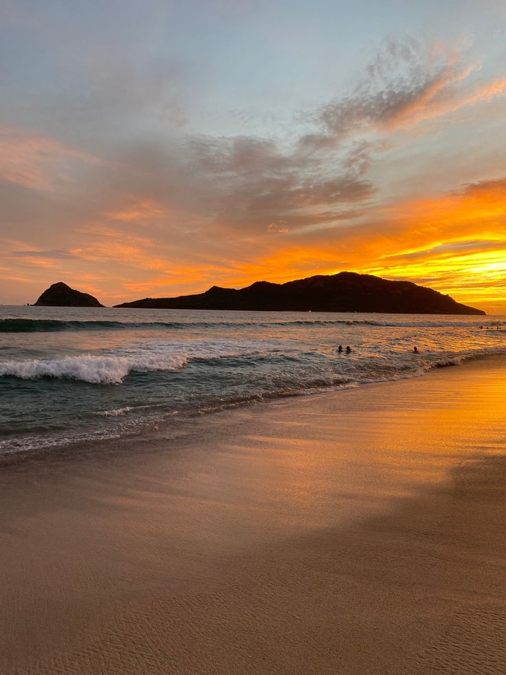 the sun is setting at the beach with people swimming in the water and hills in the distance