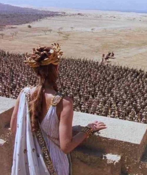 a woman standing on top of a stone wall next to a field of men in armor