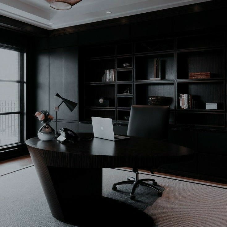 an office with black walls and flooring is lit by the window, along with a desk that has a laptop on it