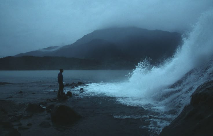 a man standing on top of a rocky beach next to a large wave in the ocean