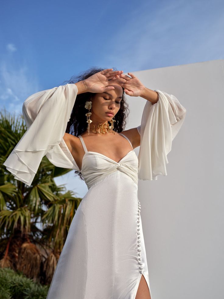 a woman in a white dress is holding her hands up to her head and looking off into the distance