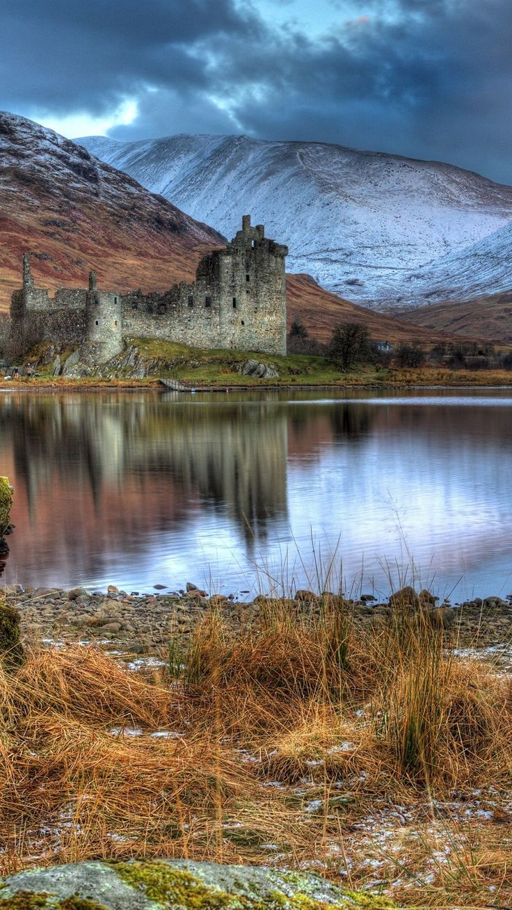 an old castle sitting on top of a mountain next to a lake with snow covered mountains in the background