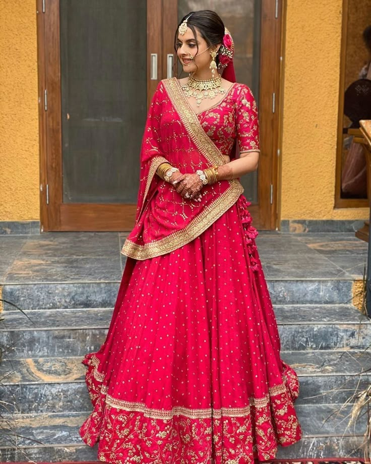 a woman in a red and gold lehenga standing on steps with her hands behind her back