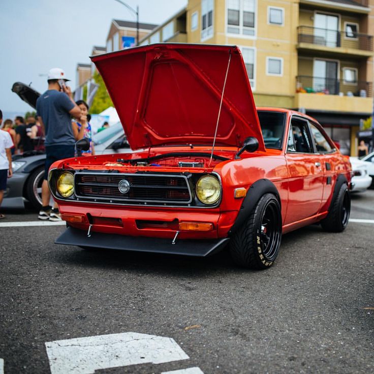 an orange car with its hood open on the street in front of some people and buildings