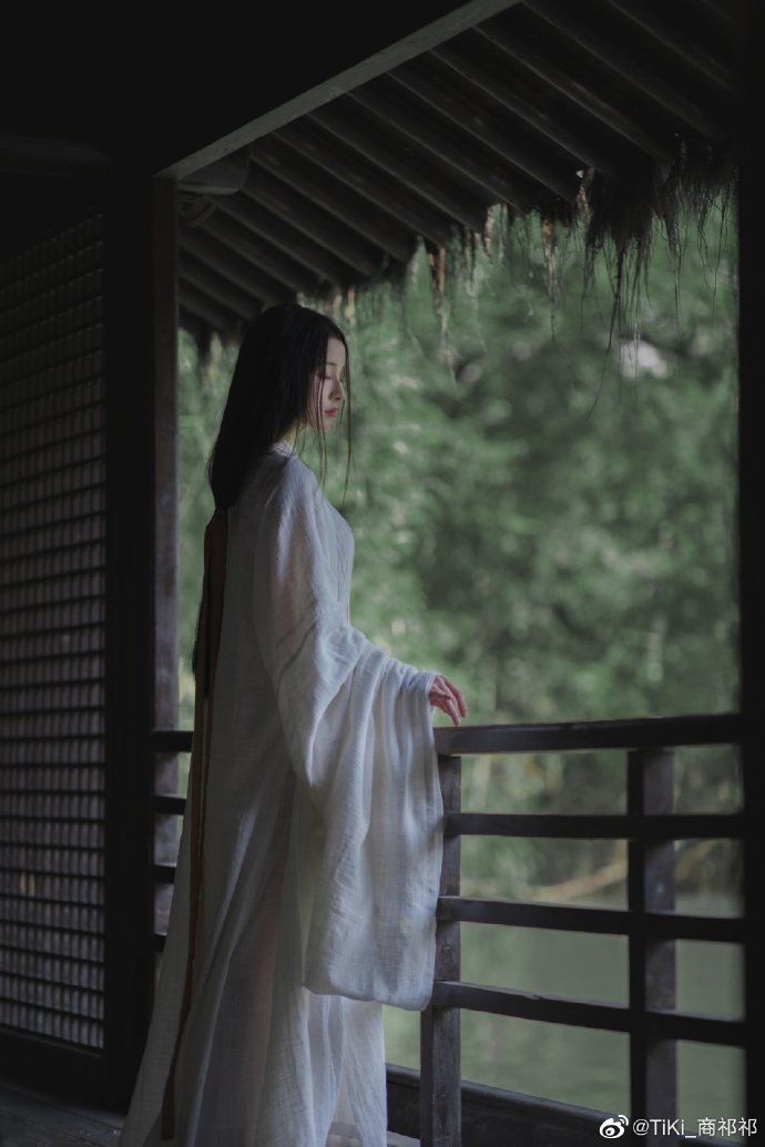 a woman dressed in white standing on a porch next to a tree and water area