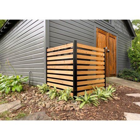 a wooden fence in front of a gray building with green plants and rocks on the ground