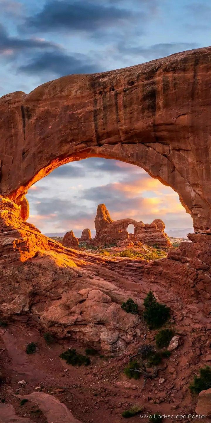 an arch shaped rock formation with the sun setting in it's center and clouds overhead