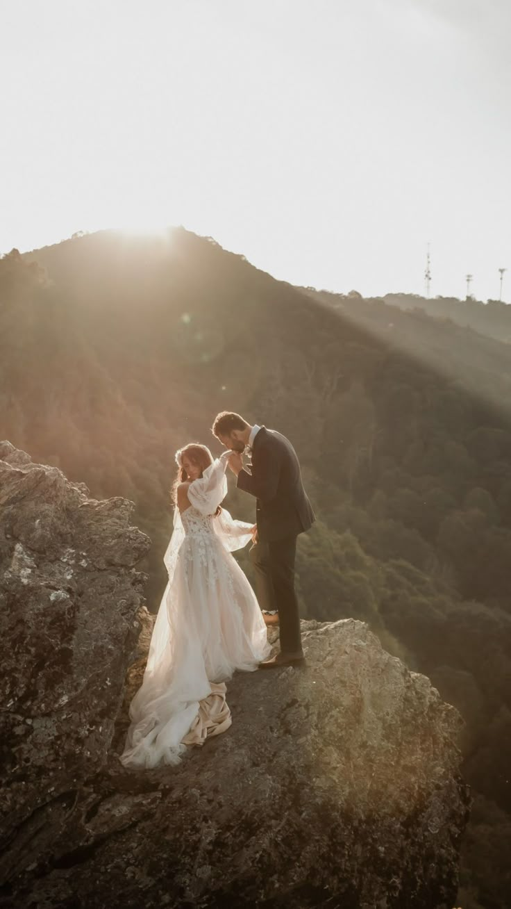 a bride and groom standing on top of a rocky hill with the sun behind them