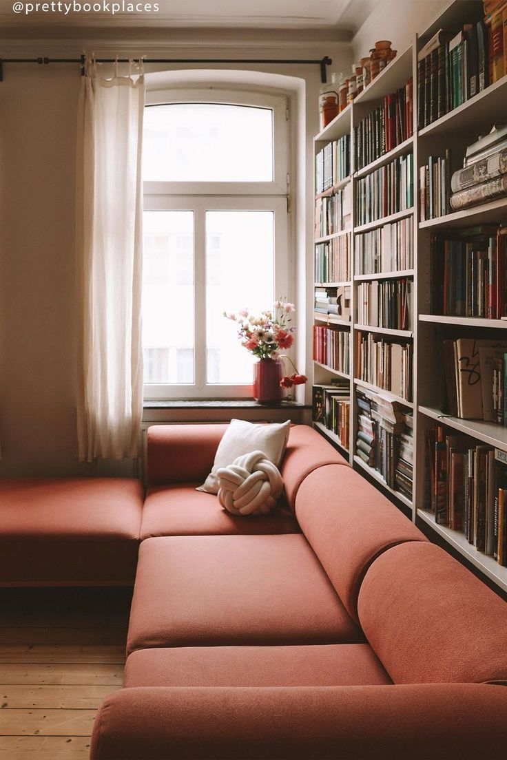 a red couch sitting in front of a book shelf filled with lots of books next to a window