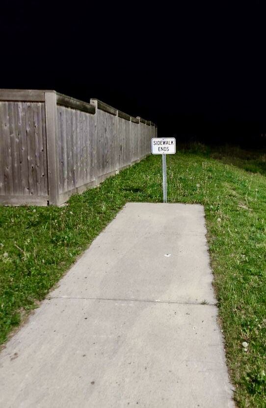 a white sign sitting on the side of a sidewalk next to a wooden fence at night