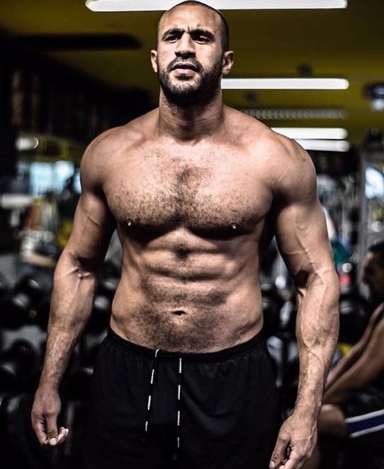 a man without a shirt standing in front of a gym machine with his hands on his hips