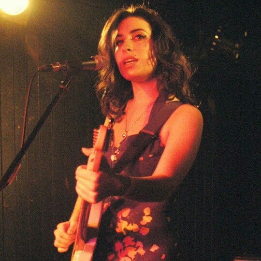 a woman standing in front of a microphone and holding a guitar while wearing a dress