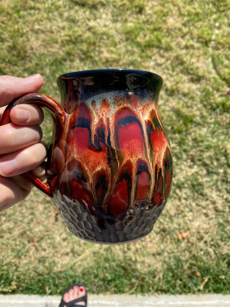 a hand holding a coffee mug with red and black swirls on the outside, in front of grass