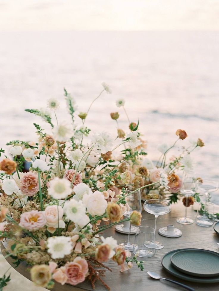 a table with flowers and wine glasses on it near the water's edge at sunset