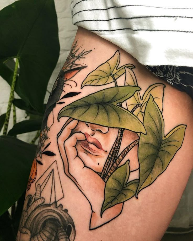 a woman's thigh with tattoos and leaves on it, showing her face behind the plant