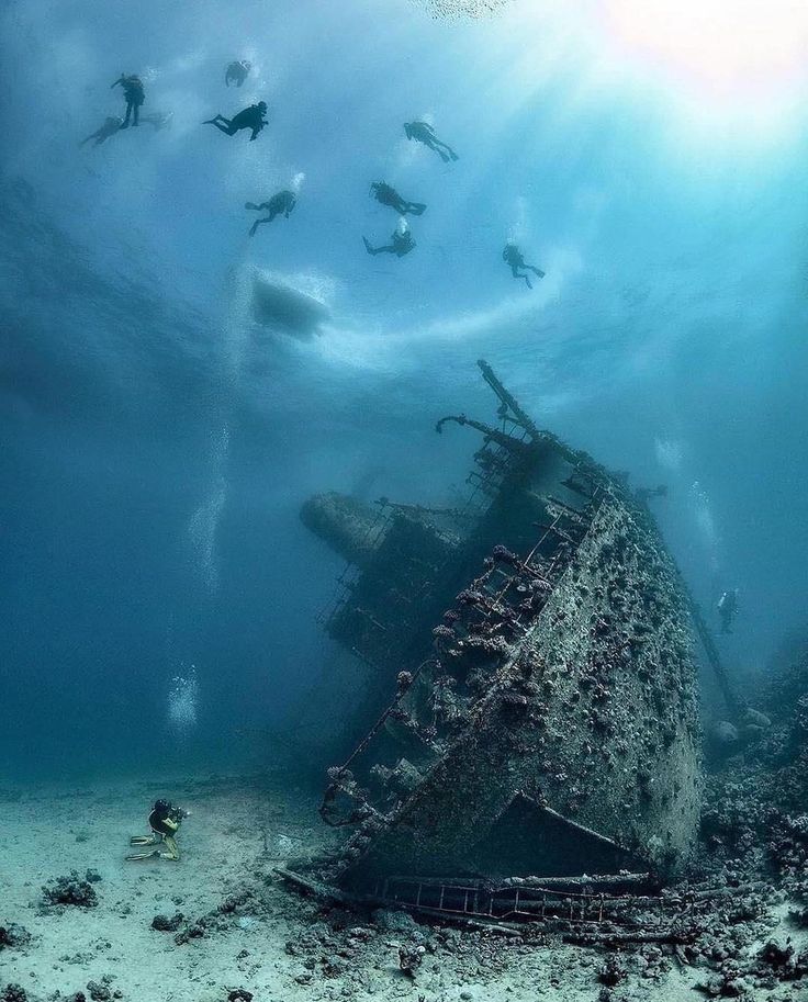 an old ship in the middle of the ocean with scubas around it and people swimming nearby