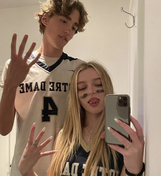 a girl with her eyes closed standing next to a boy in a basketball jersey and holding a cell phone