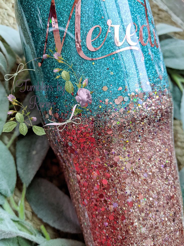 a blue and red glittered cup sitting on top of green leaves with the word merly painted on it