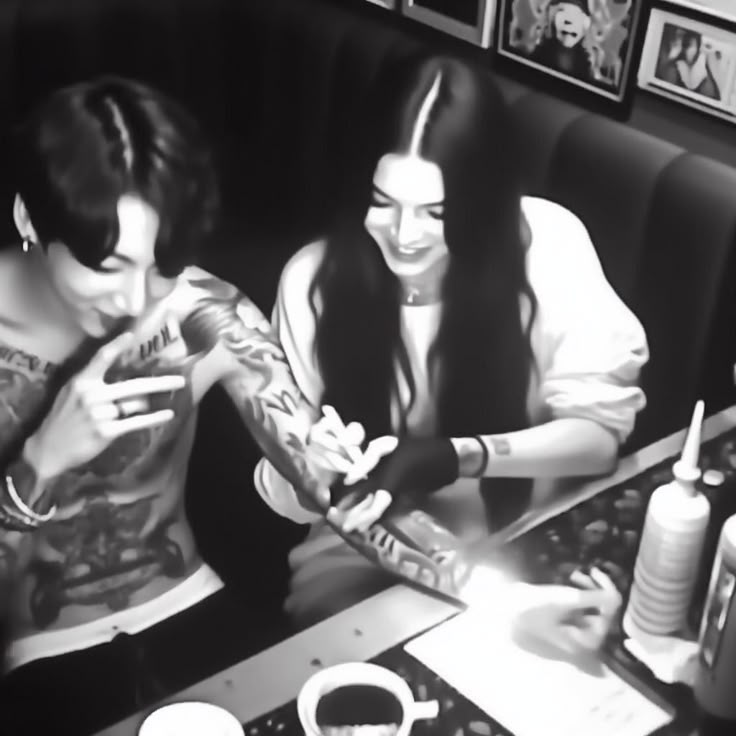 two people sitting at a table with tattoos on their arms and hands, one holding a cell phone