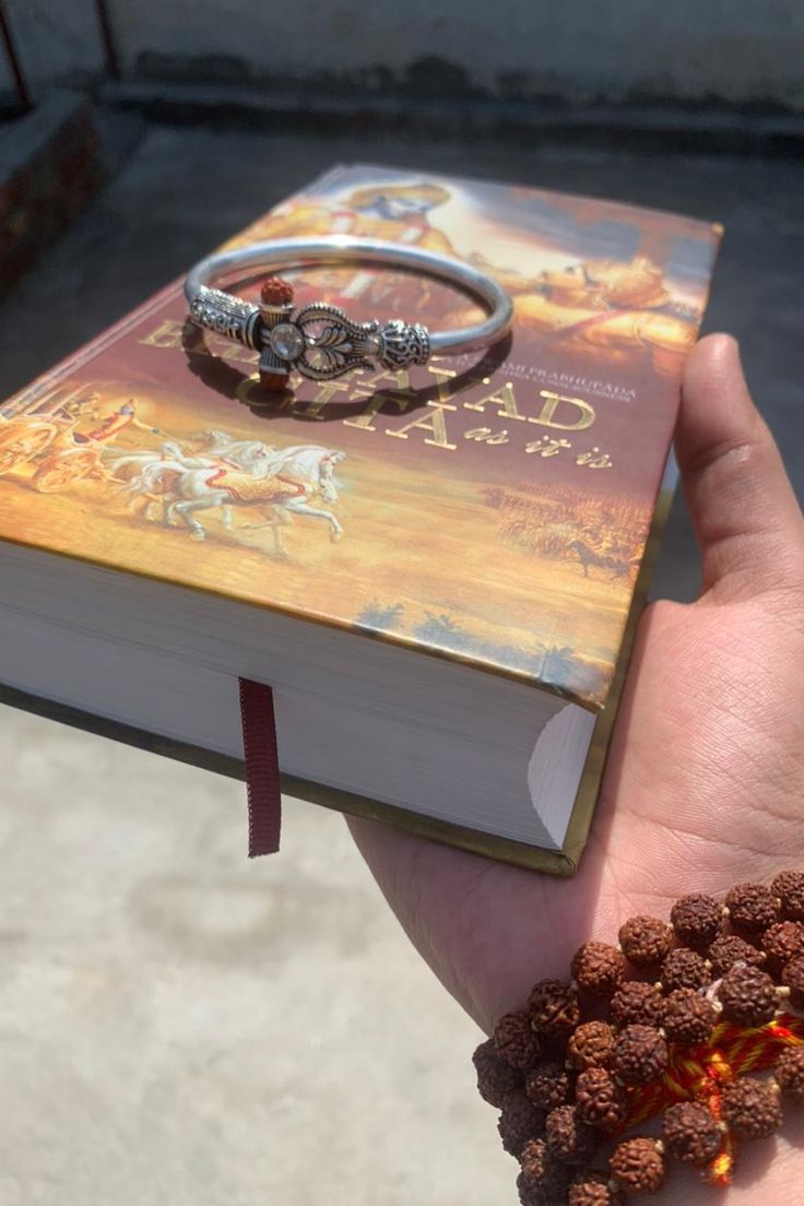 a hand holding a book with a ring on it next to a beaded bracelet