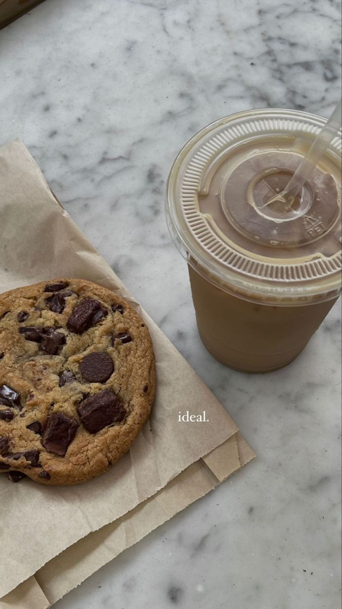 a chocolate chip cookie next to a cup of coffee on a marble counter top with a brown paper bag