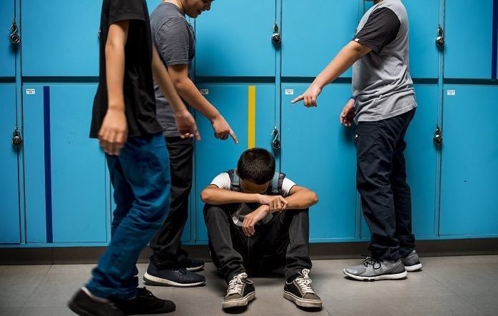 three young men standing in front of lockers with one sitting on the floor and looking at his cell phone