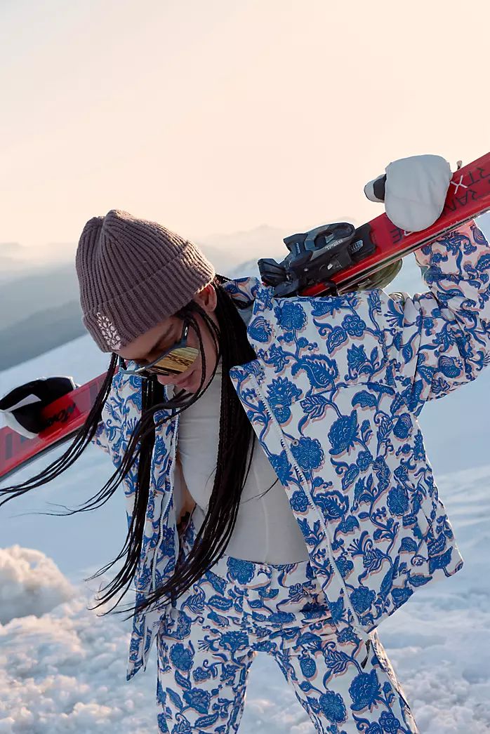 a woman in blue and white snow suit carrying skis on her back while walking through the snow