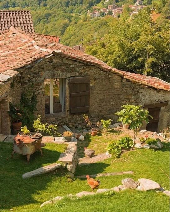 an old stone house in the middle of a grassy area with rocks and plants around it