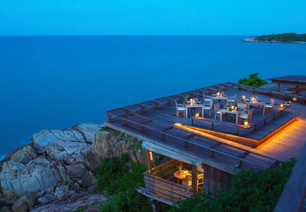 an outdoor dining area overlooking the ocean at night with lit tables and chairs on wooden planks