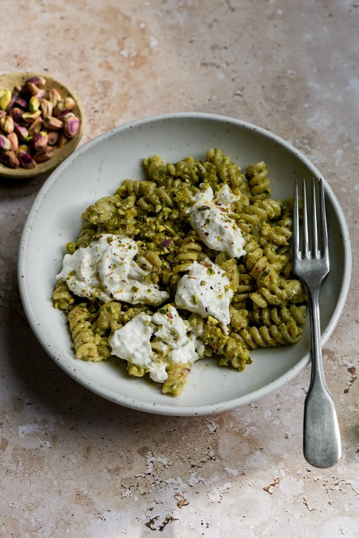 a white bowl filled with green pasta and topped with sour cream sauce next to a small bowl of pistachio seeds