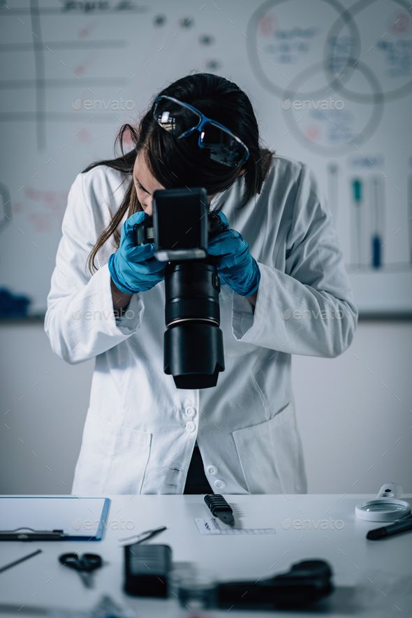 a woman in white lab coat and blue gloves holding up a camera to her face