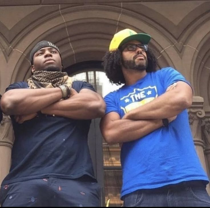 two men standing next to each other in front of a building with their arms crossed