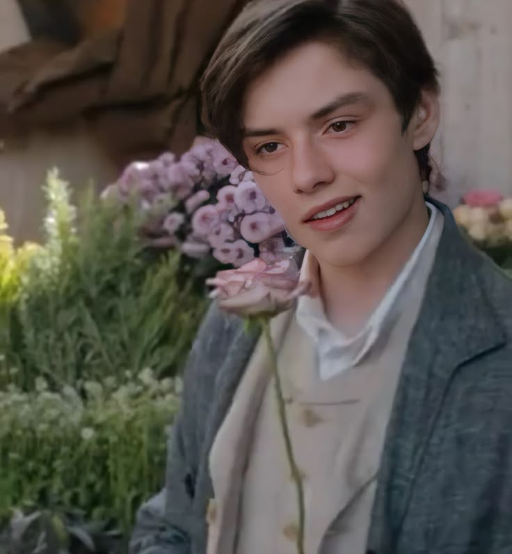 a young man holding a flower in his right hand and looking at the camera while standing next to some flowers