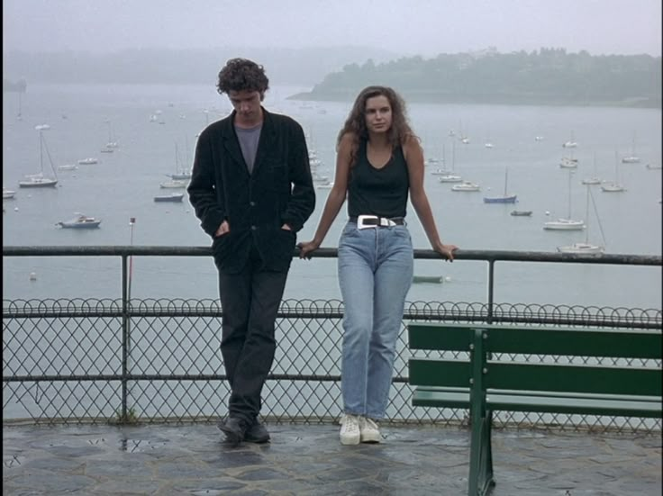 a man and woman standing next to each other on top of a stone walkway near the water